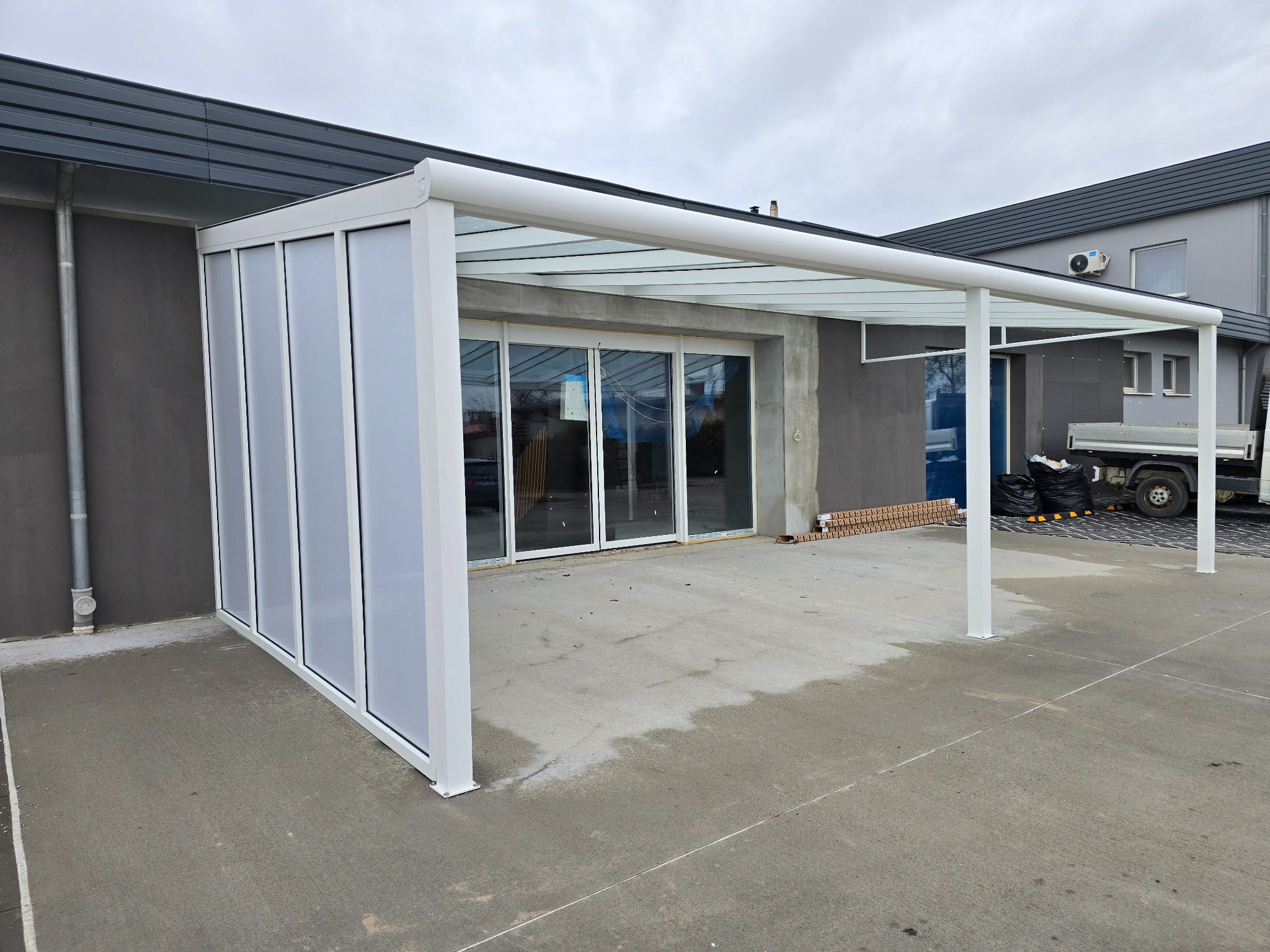 Nowoczesna, biała konstrukcja z przezroczystym zadaszeniem, częściowo zabudowana, przylegająca do budynku. Widoczny fragment elewacji i zaparkowany samochód dostawczy.