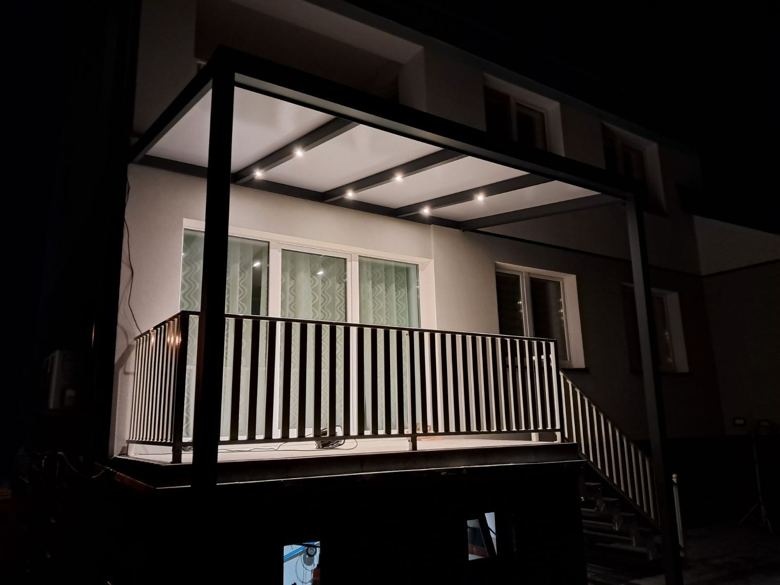 Nowoczesna, podświetlana pergola metalowa z balustradą na balkonie, oświetlona punktowymi lampami, widoczna nocą. Minimalistyczny design, ciemna kolorystyka konstrukcji.