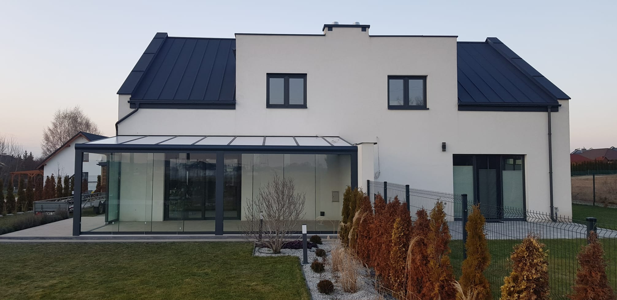 Nowoczesna, przeszklona zabudowa balkonu z ciemną ramą, przylegająca do białego domu z ciemnym dachem. Widoczne elementy ogrodu i ogrodzenie panelowe.