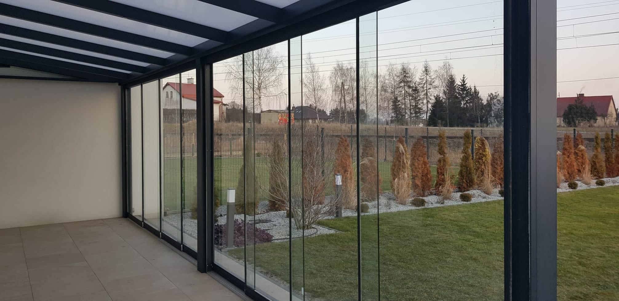 Nowoczesna zabudowa balkonu z przesuwnymi panelami szklanymi i metalową konstrukcją, widok na ogród z ozdobnymi krzewami i trawami. Minimalistyczny design.