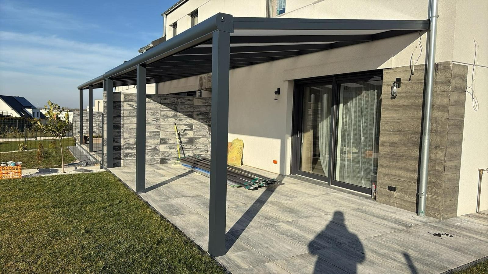 Nowoczesna, szara pergola metalowa przy domu z przesuwnymi drzwiami. Widoczne elementy konstrukcyjne i materiały przygotowane do montażu. Jasne płytki na tarasie.