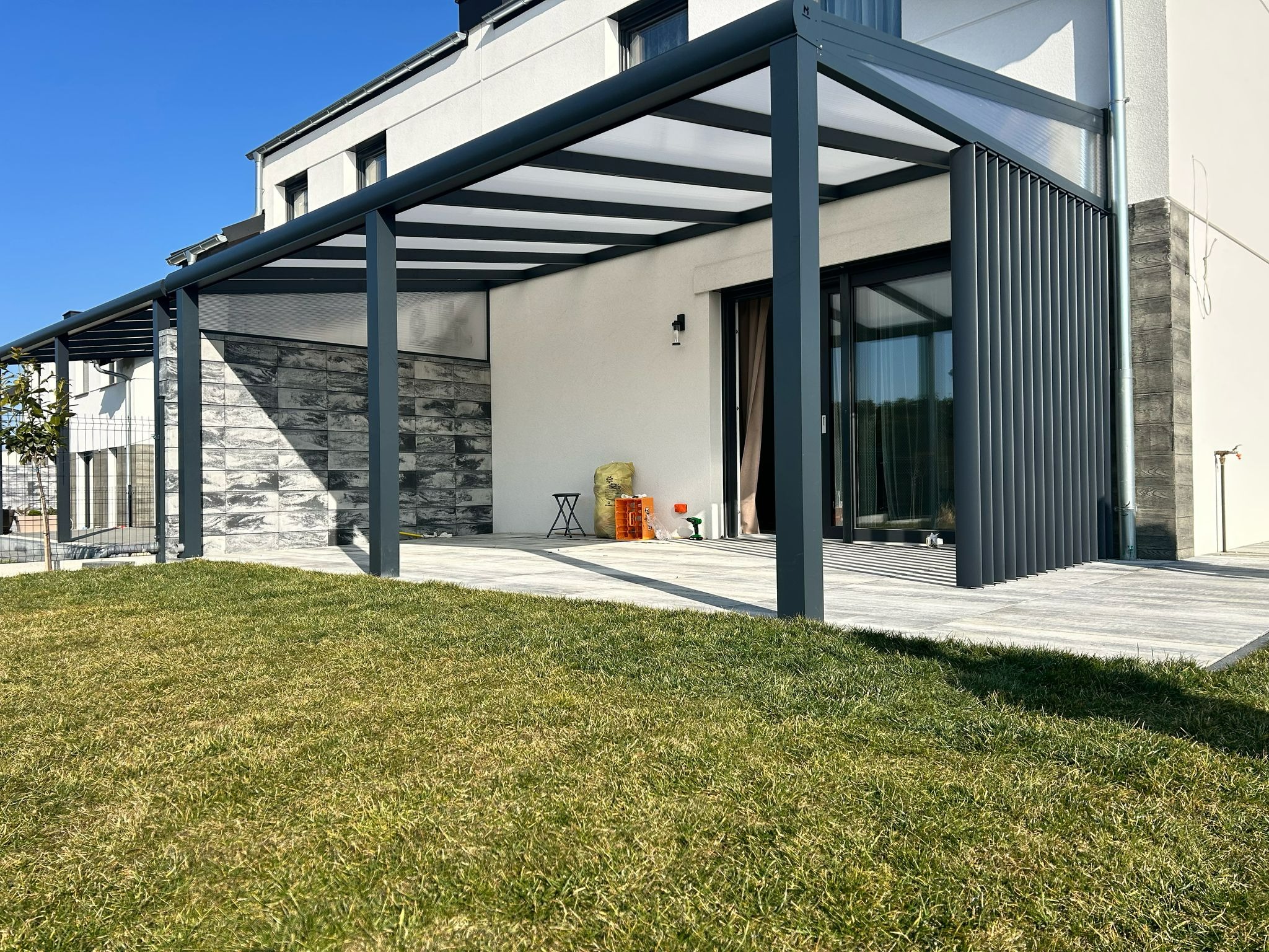 Nowoczesna pergola z szarymi metalowymi słupami i białym zadaszeniem, przylegająca do jasnego budynku z dużymi oknami, otoczona zielonym trawnikiem w słoneczny dzień.