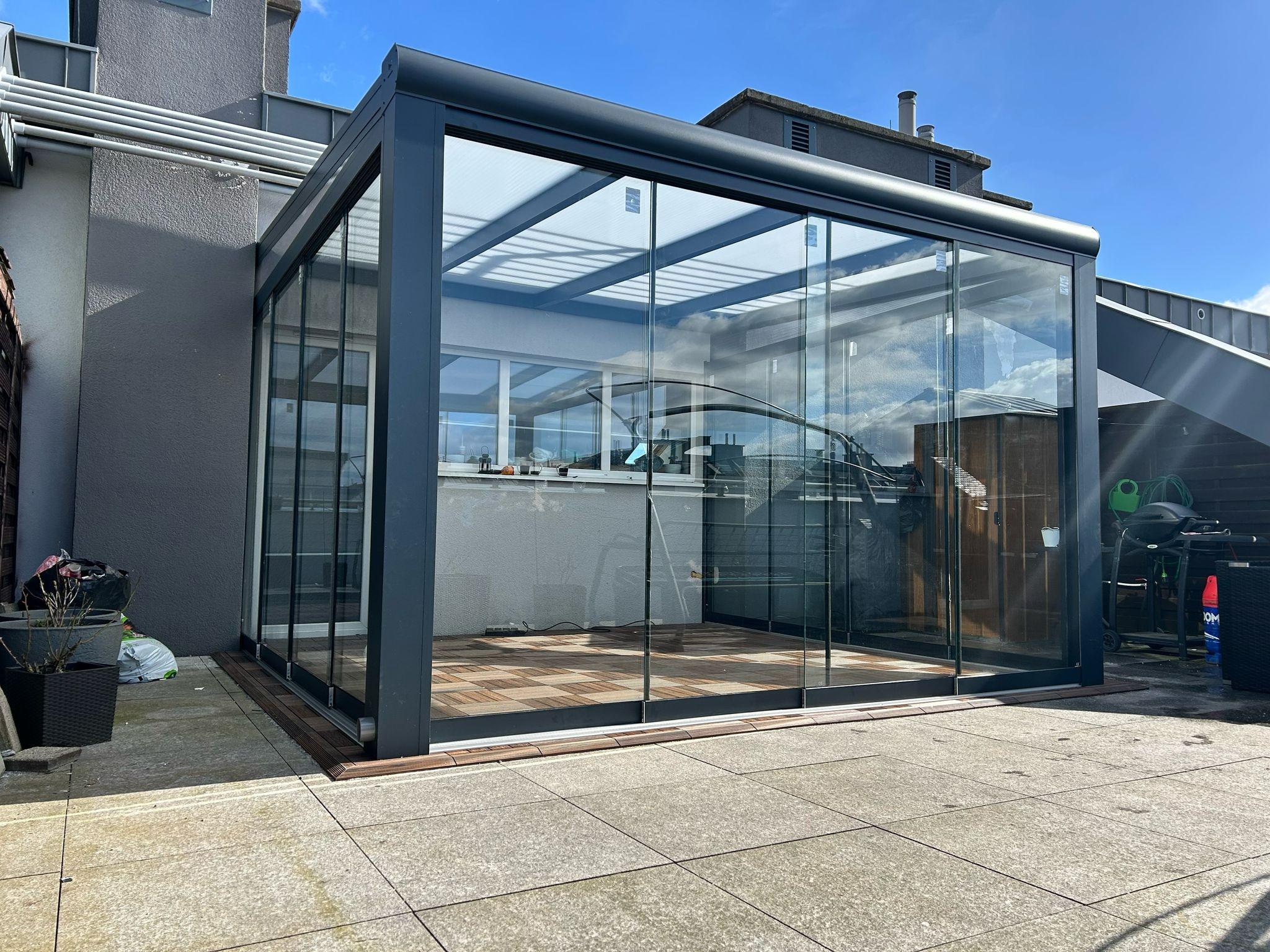 Szklana zabudowa balkonu z przesuwnymi drzwiami, widok z zewnątrz. Nowoczesna konstrukcja z ciemną ramą i drewnianą podłogą. Jasne tło nieba i budynków.