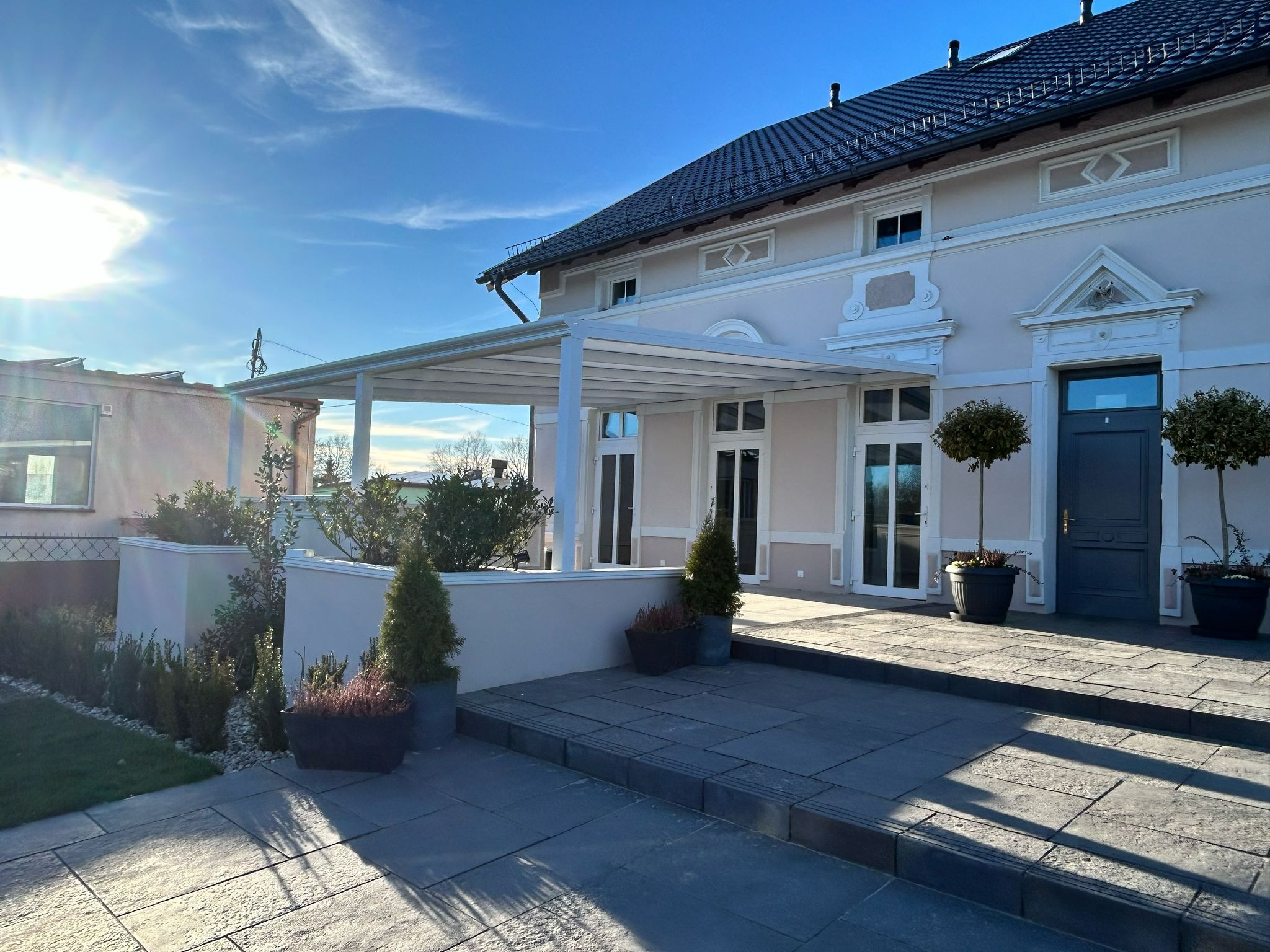Biała pergola z zadaszeniem tarasowym przylegająca do jasnego budynku z ciemnym dachem. Widoczne rośliny w donicach i kostka brukowa. Słoneczny dzień.