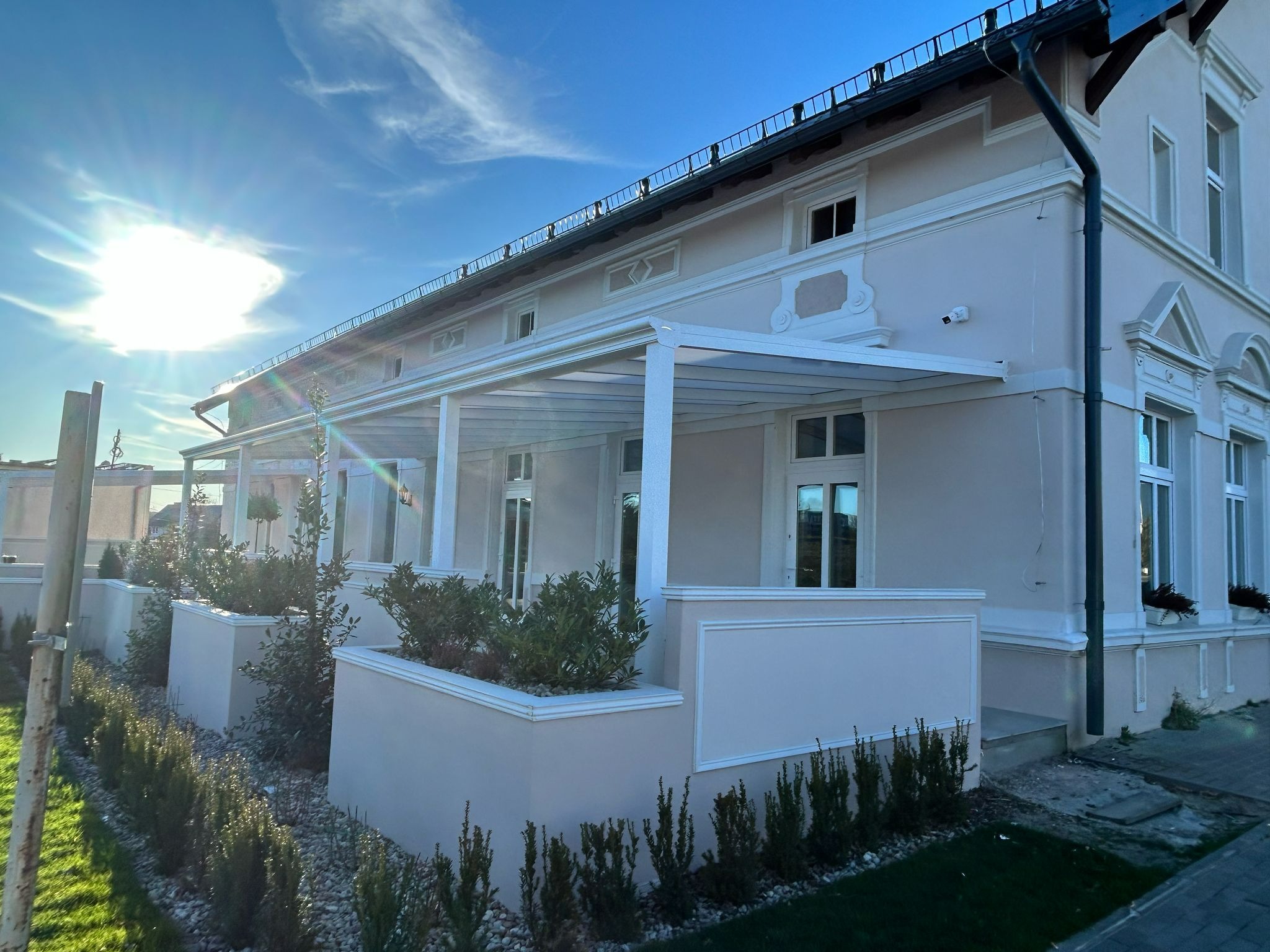 Elegancka, biała pergola metalowa przylegająca do budynku w klasycznym stylu. Widoczne rośliny w donicach i zadbany trawnik. Słoneczny dzień, błękitne niebo.