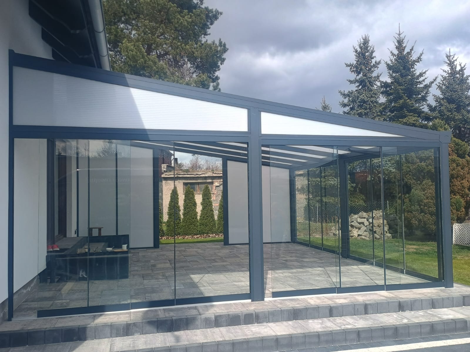 Szklana zabudowa tarasu z szarymi profilami, widok z zewnątrz. Konstrukcja zadaszona, z przesuwnymi panelami szklanymi. W tle zieleń i budynek.