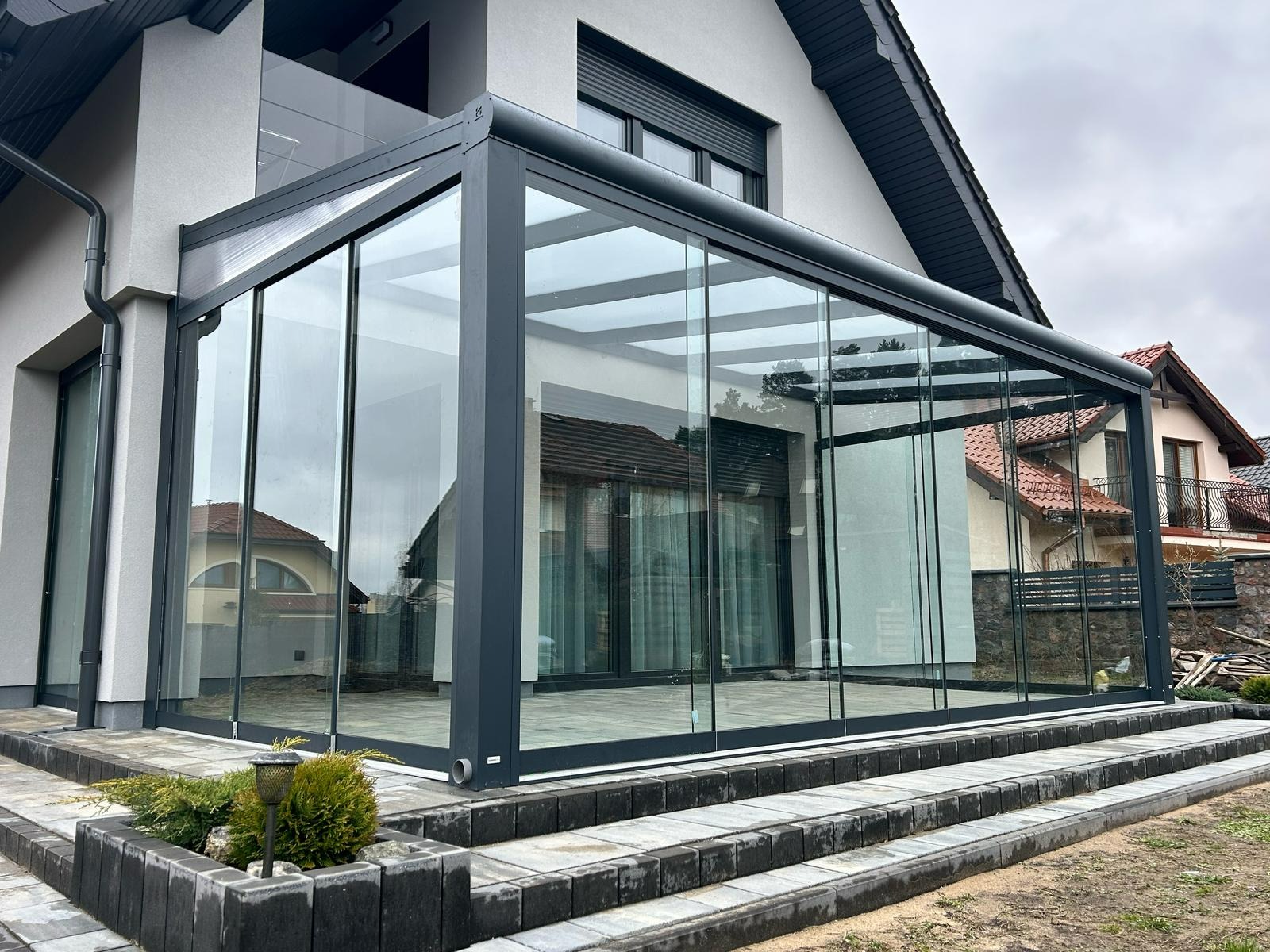 Szklana zabudowa tarasu z szarymi profilami, widoczna z zewnątrz, z niskim oświetleniem ogrodowym i schodami z kostki brukowej prowadzącymi do wejścia.