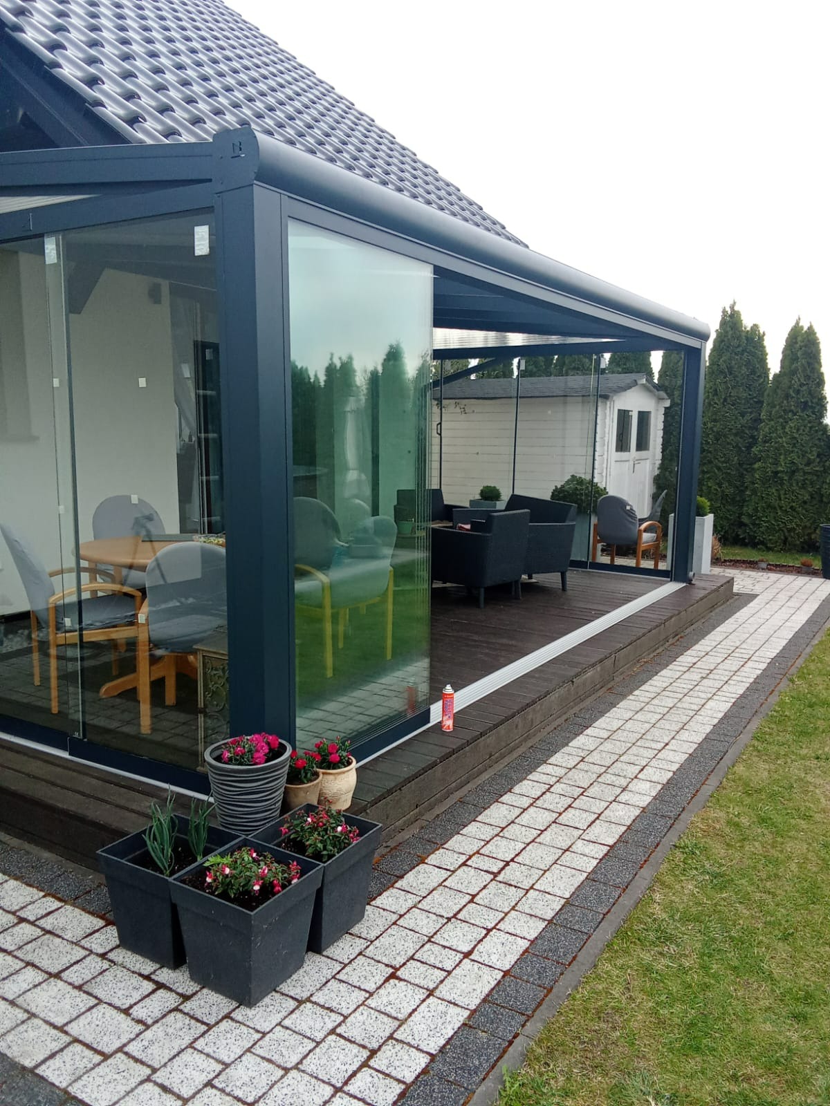 Szklana zabudowa tarasu z ciemną ramą w Dobczycach, widok z zewnątrz. Na pierwszym planie donice z kwiatami, w tle meble ogrodowe i zieleń.