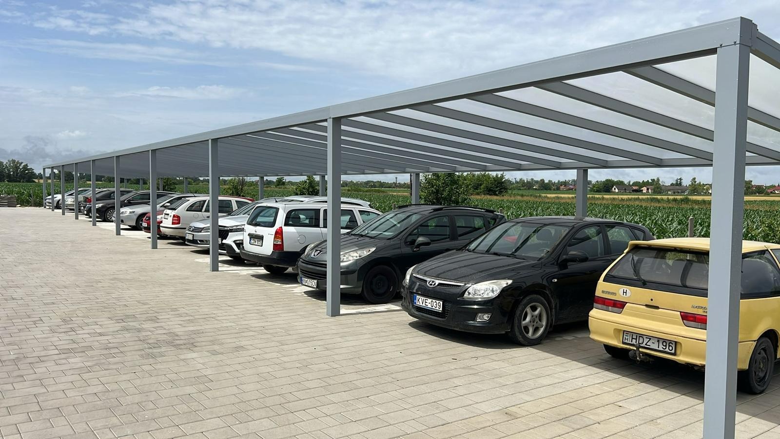 Zadaszenie parkingowe z metalową konstrukcją i przezroczystym dachem, chroniące zaparkowane samochody. Widok na pole w tle, jasne niebo.