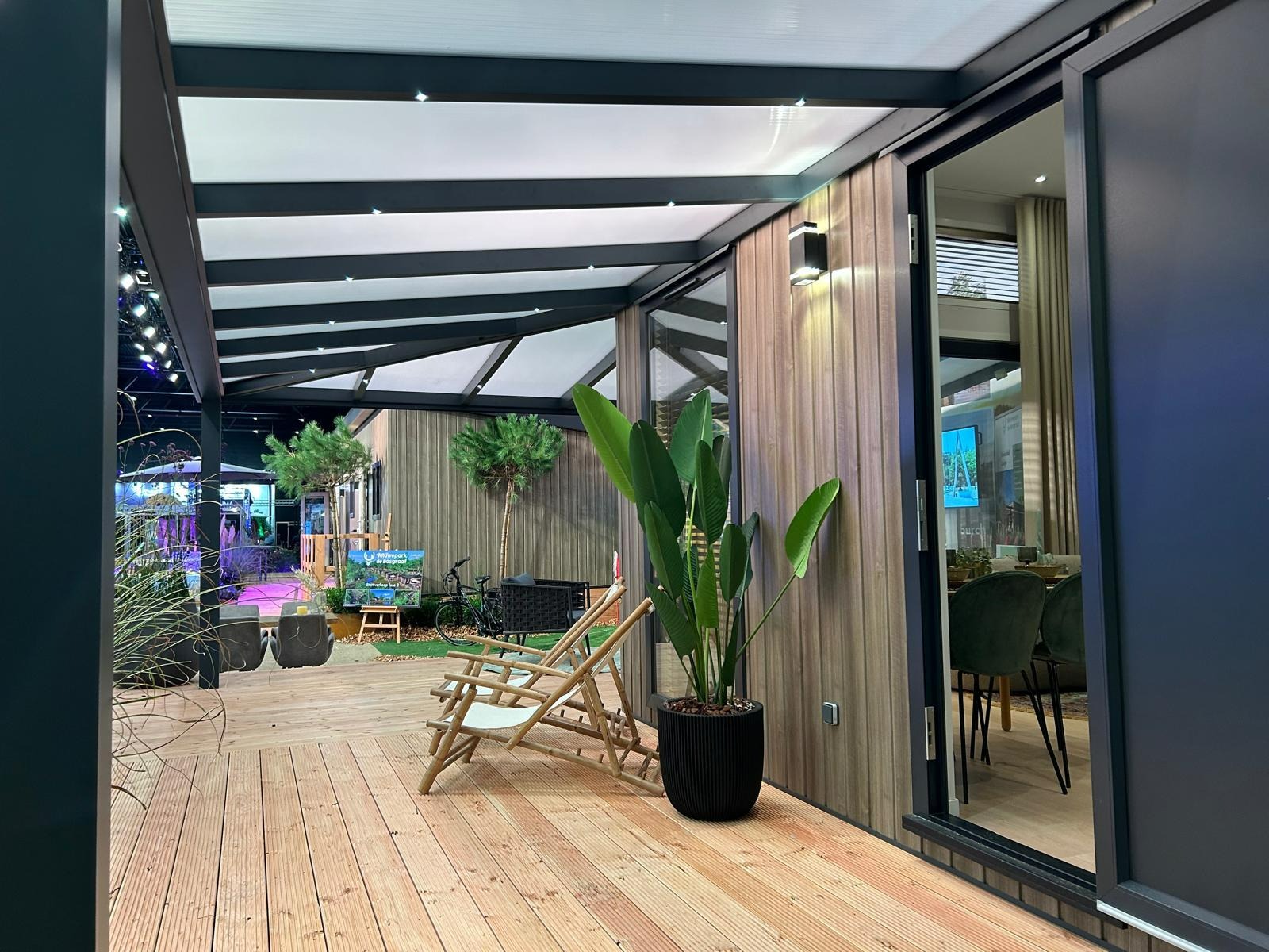 Zadaszony taras z drewnianą podłogą i meblami ogrodowymi. Widoczne elementy konstrukcji z metalu i drewniane wykończenie ścian. W tle zieleń i zabudowa.