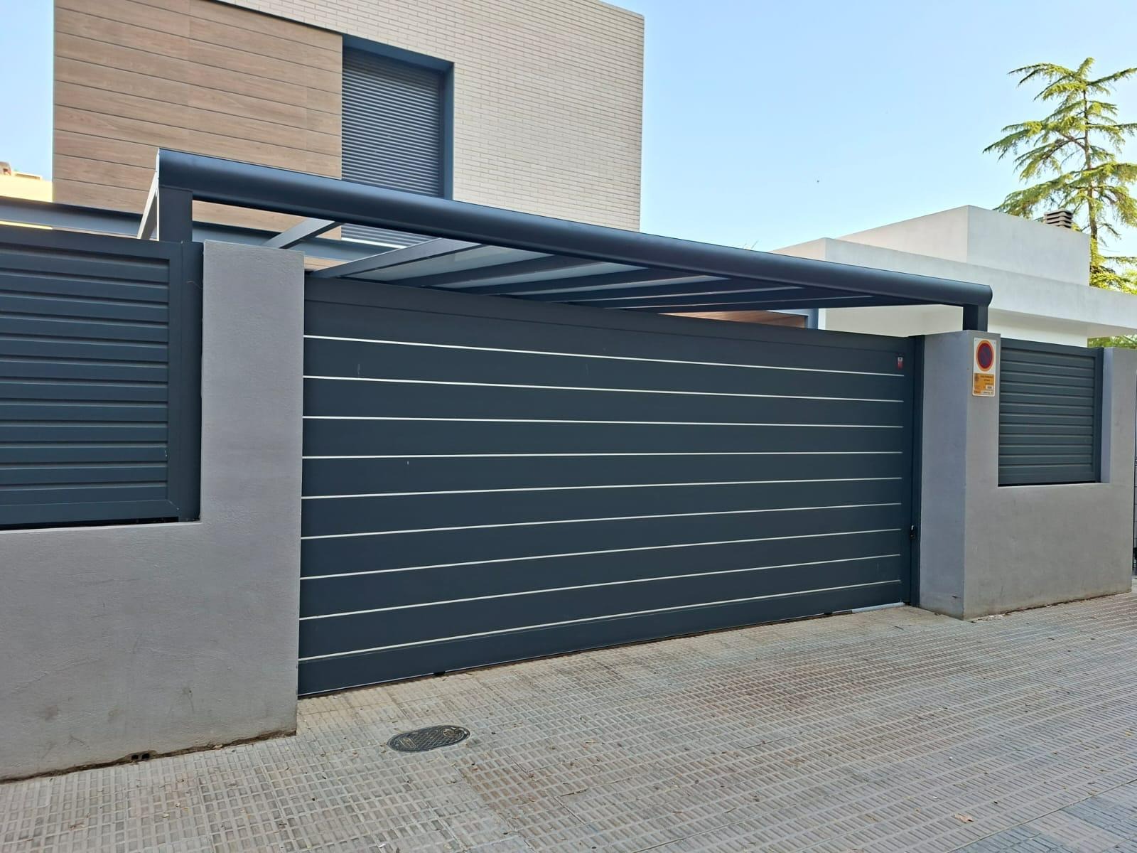 Nowoczesna, przesuwna brama wjazdowa z poziomymi panelami i zadaszeniem, w kolorze antracytowym, na tle jasnego budynku mieszkalnego. Minimalistyczny design.