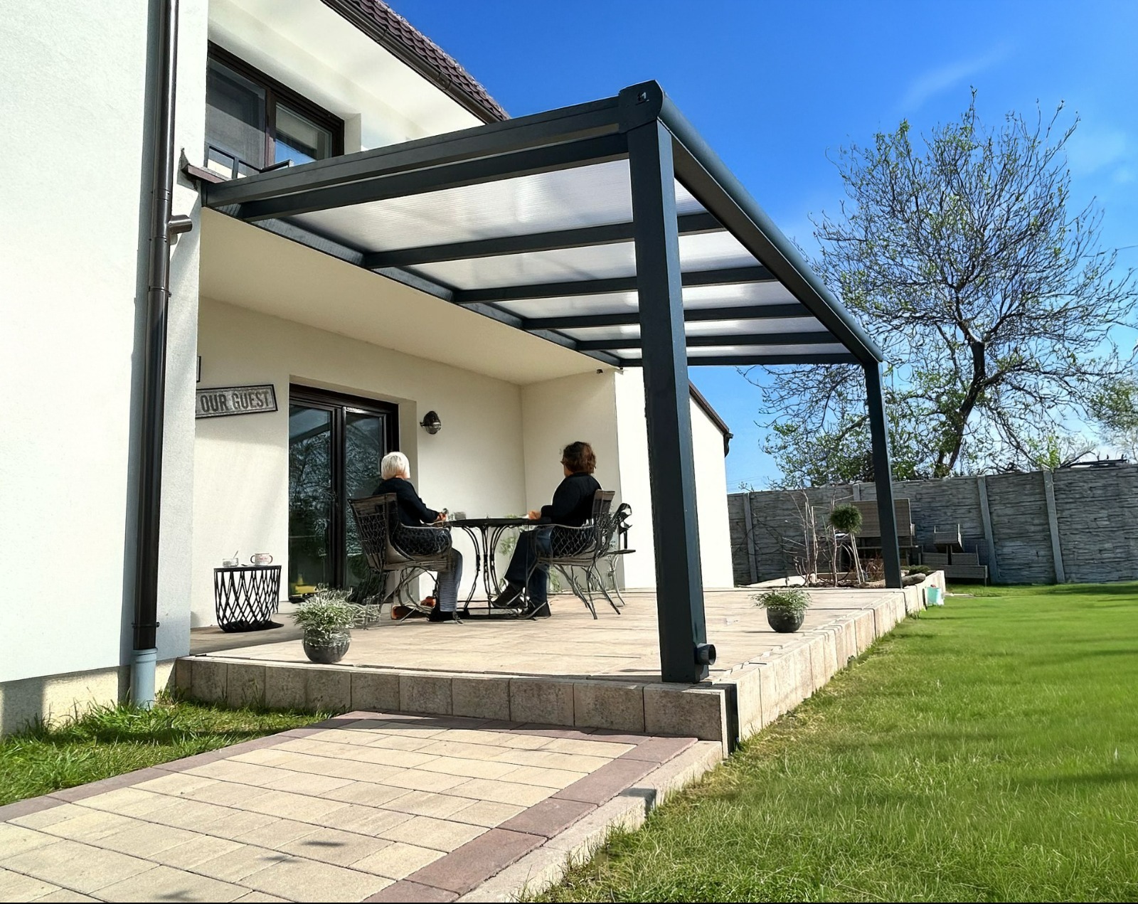 Nowoczesna, grafitowa pergola metalowa z przezroczystym zadaszeniem, przylegająca do białego domu. Dwie osoby siedzą przy stoliku na tarasie. Widok na ogród i błękitne niebo.