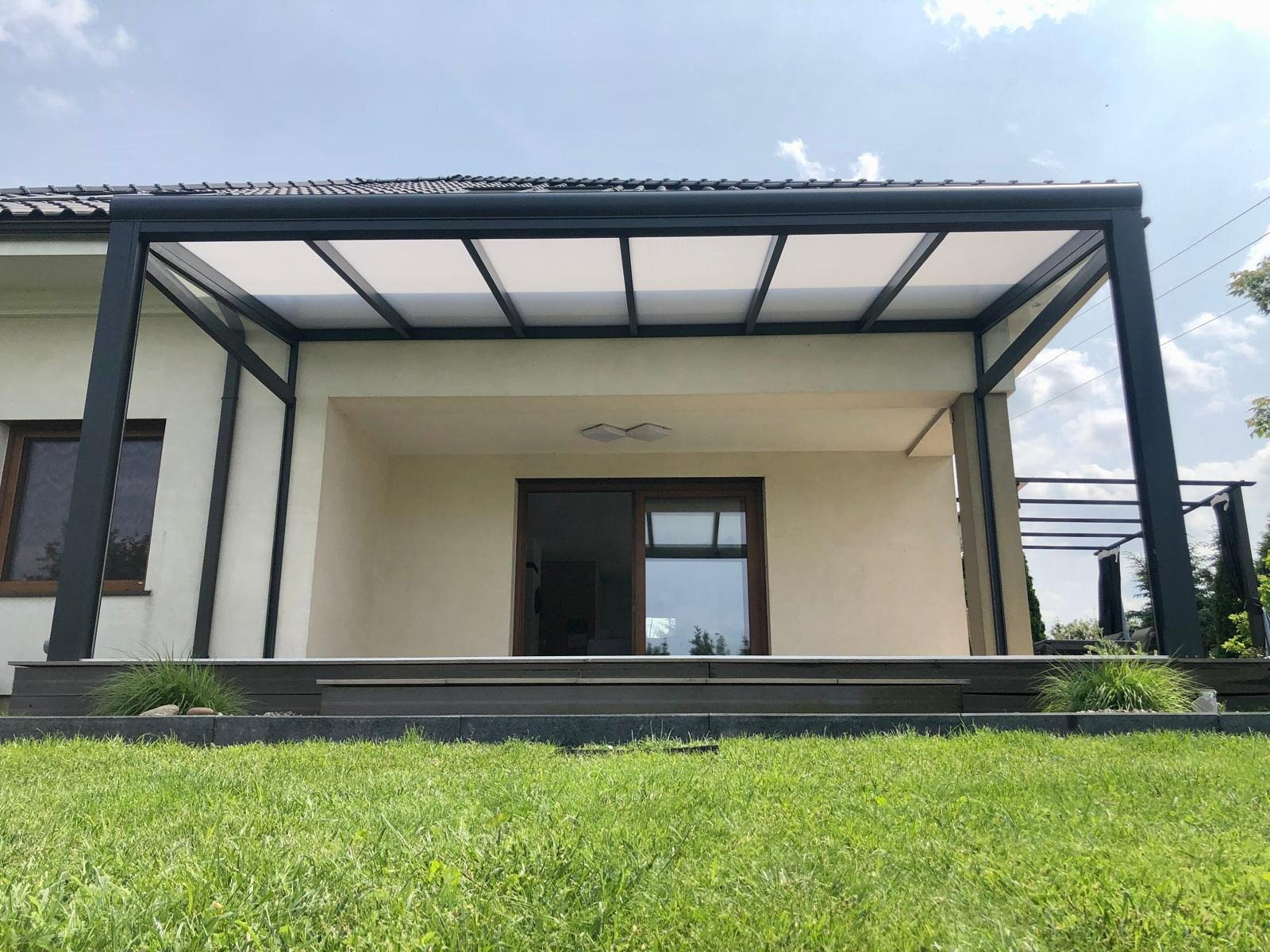Nowoczesna pergola z metalową konstrukcją i zadaszeniem z paneli, przylegająca do budynku. Widok z poziomu trawnika, zieleń w kadrze. Minimalistyczny design.