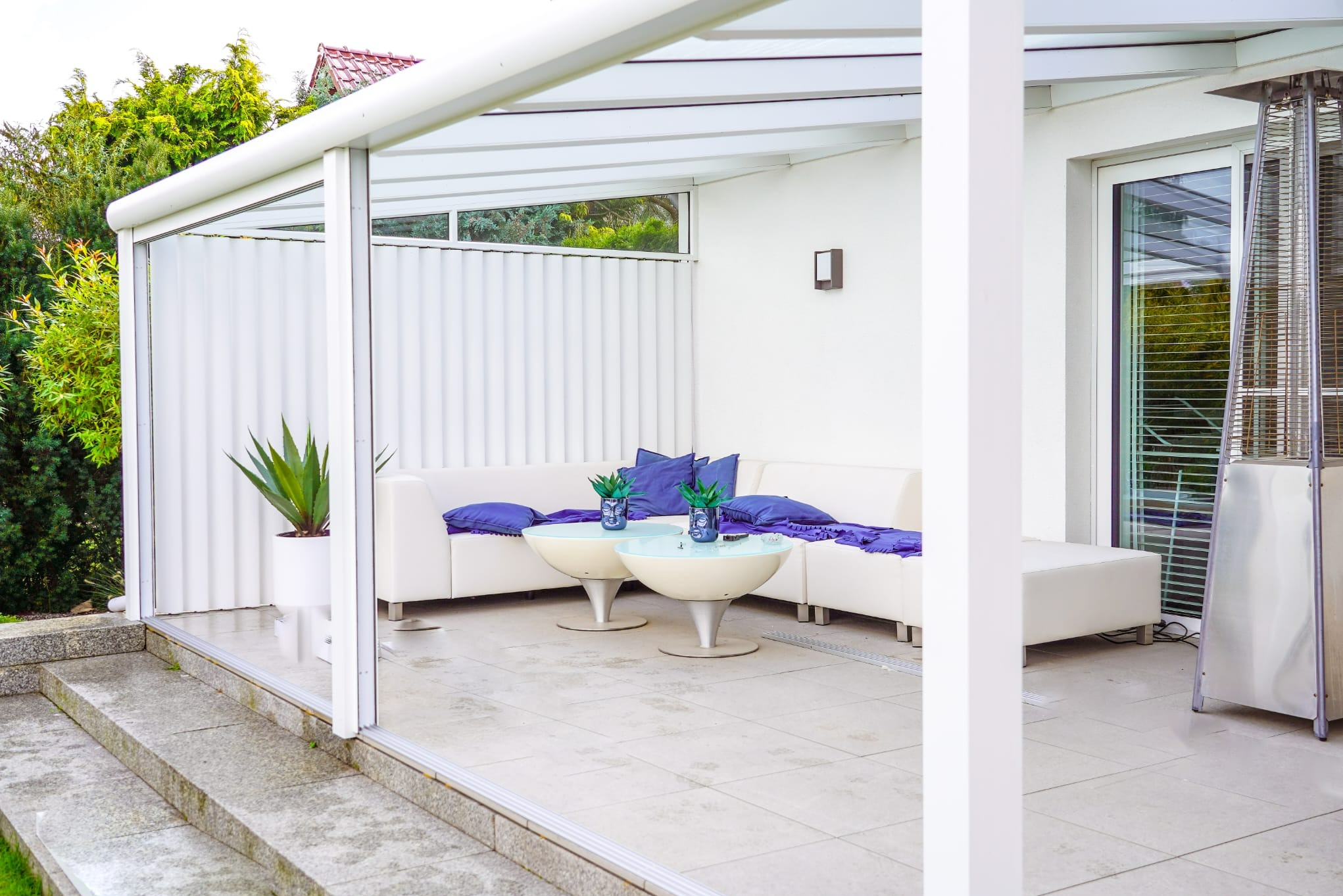 Nowoczesna, biała pergola z metalową konstrukcją i szklanym dachem, tworząca komfortową strefę relaksu z sofami i stolikami na tarasie.