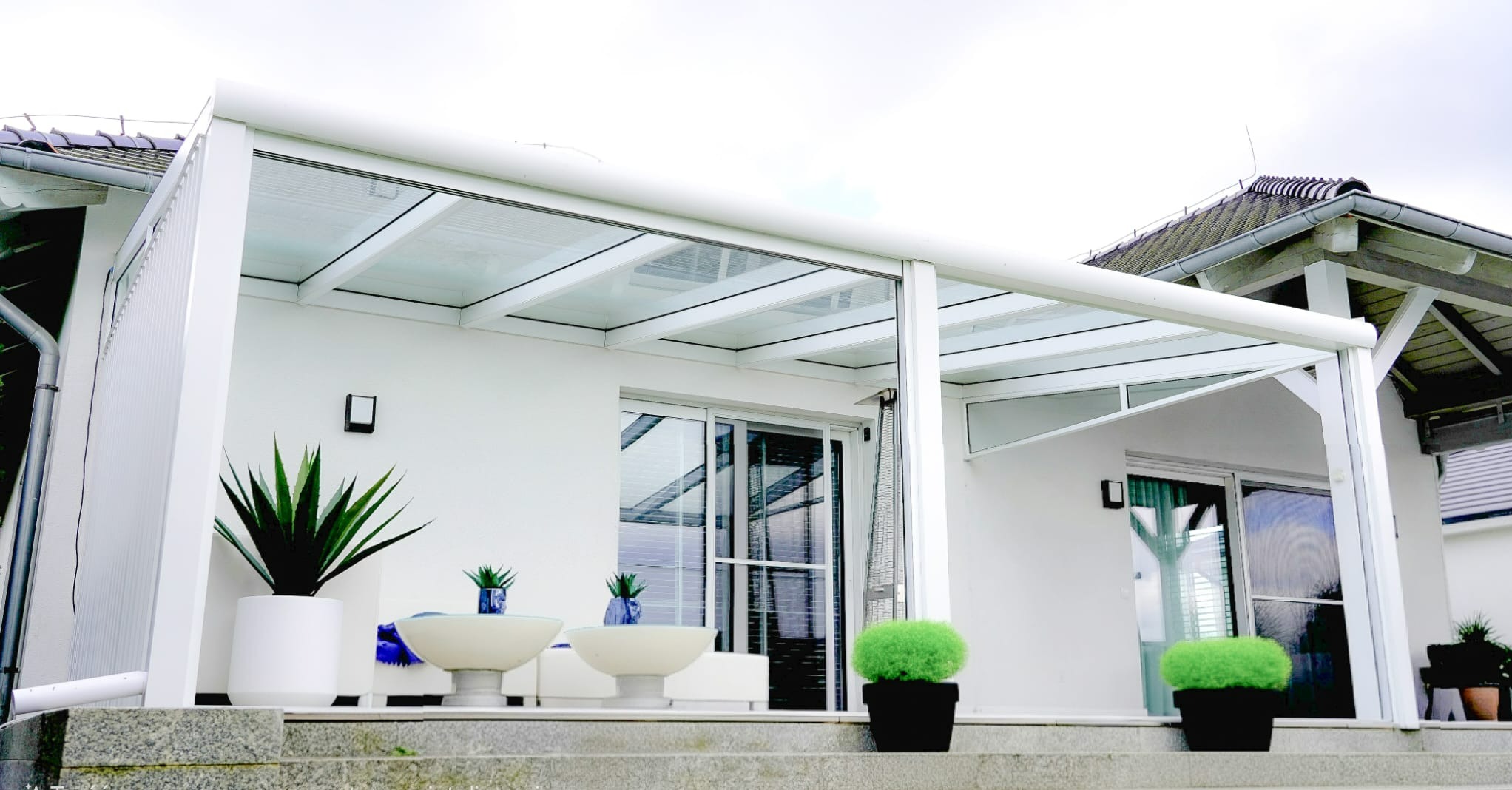 Biała, nowoczesna pergola z przezroczystym dachem, osłaniająca taras z roślinami w donicach. Widok z dołu, podkreślający minimalistyczny design i funkcjonalność.