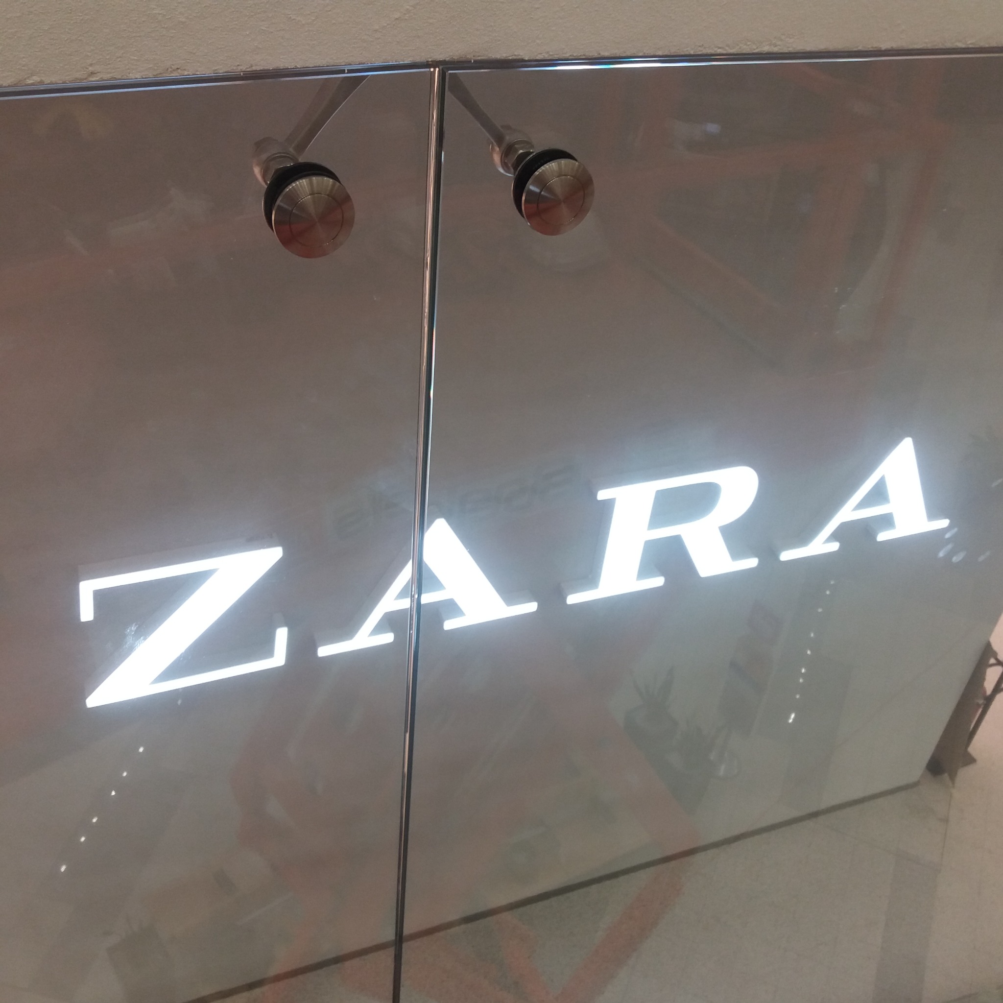Szklane drzwi z podświetlonym logo 'ZARA', widoczne metalowe okucia. Ujęcie z dołu, perspektywa podkreśla wielkość logo. Minimalistyczny design.