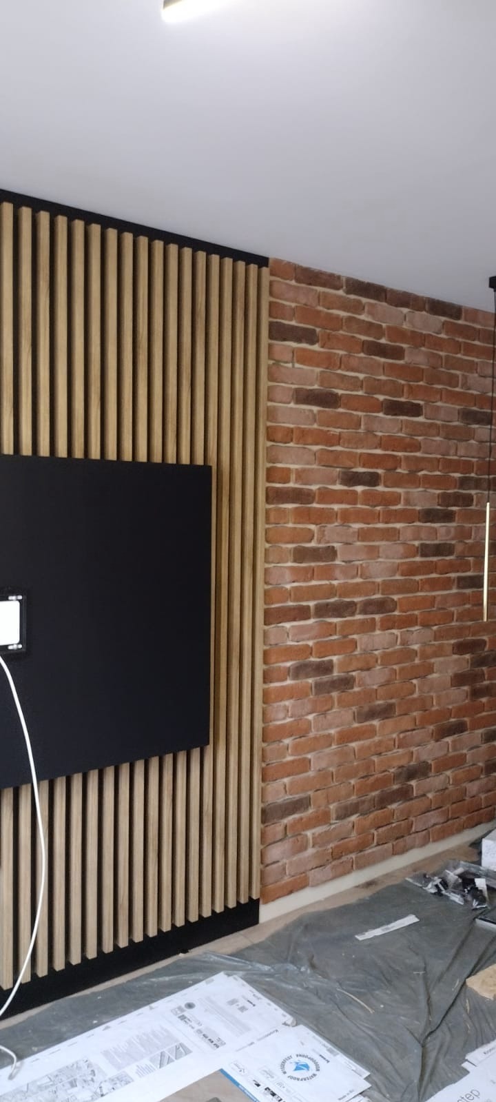 Nowoczesna ściana z dekoracyjną cegłą i drewnianymi lamelami, na której wisi czarny telewizor. Widoczne materiały budowlane na podłodze.