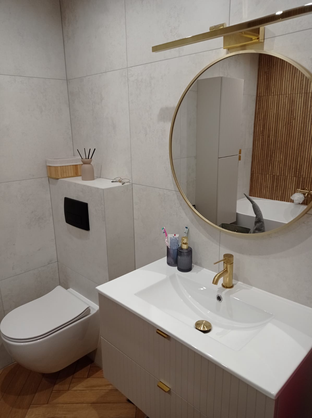 Nowoczesna łazienka w Czernikowie: białe płytki, wisząca toaleta, umywalka z szafką i okrągłe lustro z oświetleniem. Widoczne złote akcenty.