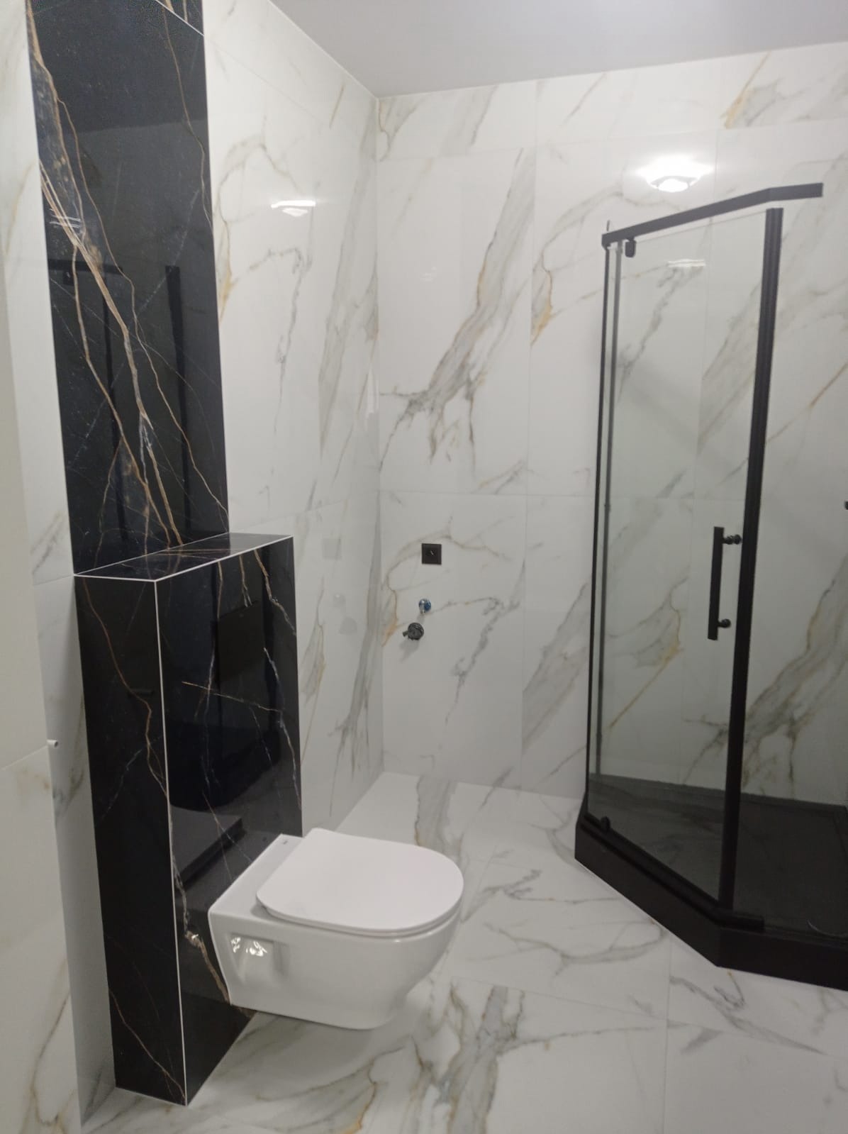 Elegancka łazienka z kabiną prysznicową i podwieszaną toaletą. Płytki imitujące marmur z czarnymi akcentami. Nowoczesny design, minimalistyczne baterie.