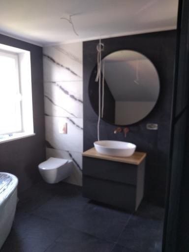 Nowoczesna łazienka z okrągłym lustrem, umywalką nablatową i płytkami imitującymi marmur. Widoczne elementy instalacji elektrycznej.