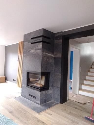 Nowoczesny kominek obłożony szarym kamieniem z czarnymi akcentami, drewniana podłoga, widok na schody i drzwi w tle. Minimalistyczny design.