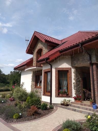 Elewacja domu z czerwonym dachem i kamiennymi elementami. Okna z ceglaną obudową, zadbany ogród i ścieżka z kostki brukowej. Ujęcie w słoneczny dzień.