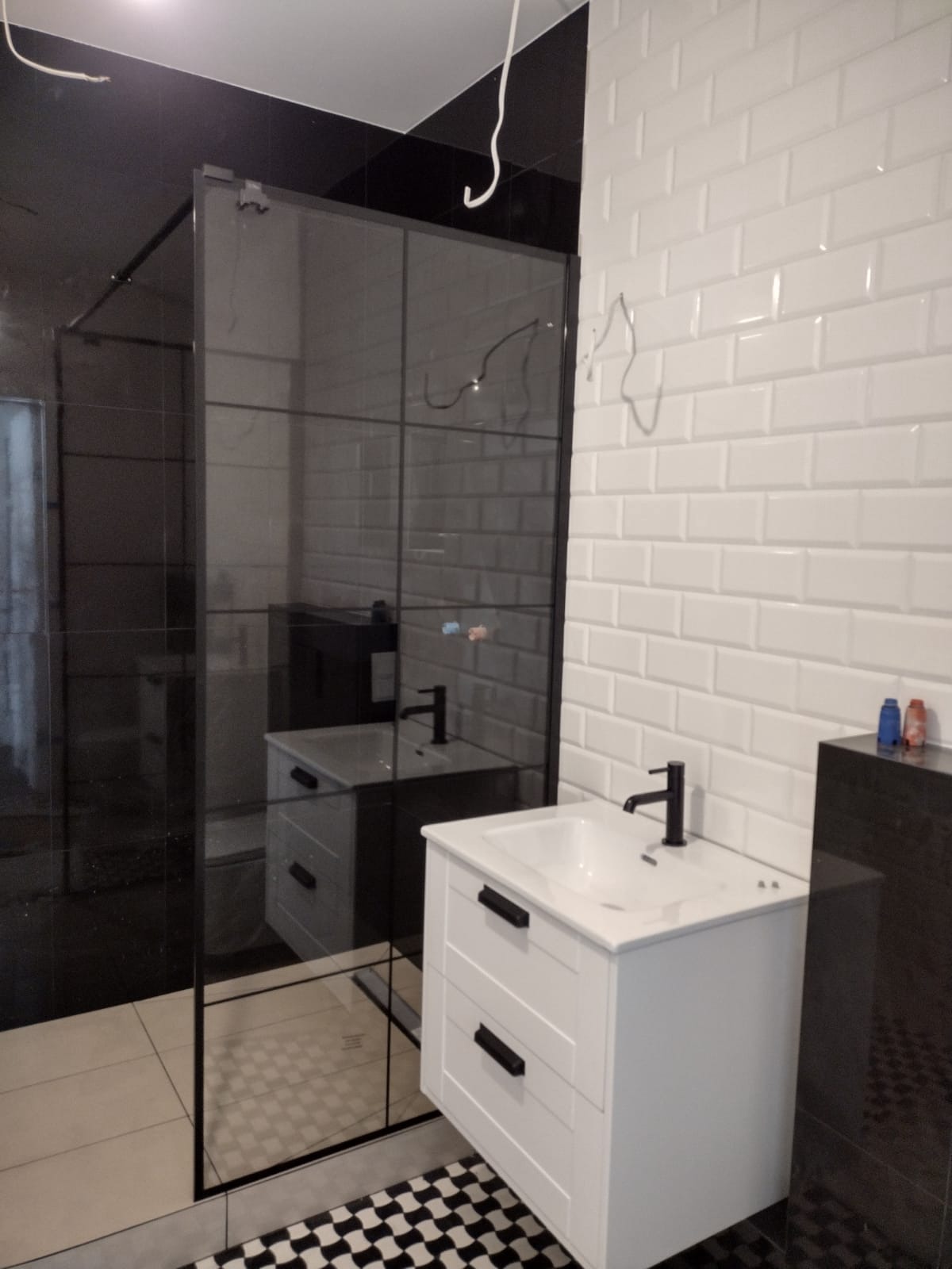 Nowoczesna łazienka z kabiną prysznicową typu walk-in, białą umywalką z szafką i białą cegłą na ścianie. Czarno-biała szachownica na podłodze dodaje kontrastu.