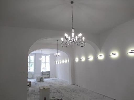 Białe wnętrze po malowaniu z żyrandolami i oświetleniem ściennym. Podłoga zabezpieczona folią malarską. Widok przez łukowate przejście do kolejnego pomieszczenia.