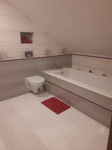 Biała łazienka na poddaszu z wanną, toaletą podwieszaną i czerwonym akcentem w postaci dywanika i listwy dekoracyjnej na płytkach.