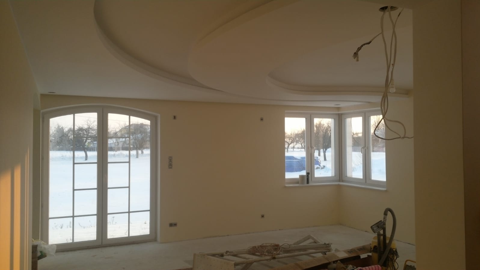 Jasne wnętrze po szpachlowaniu ścian i sufitu z dekoracyjną sztukaterią. Widoczne okna z widokiem na zimowy krajobraz i narzędzia na podłodze.