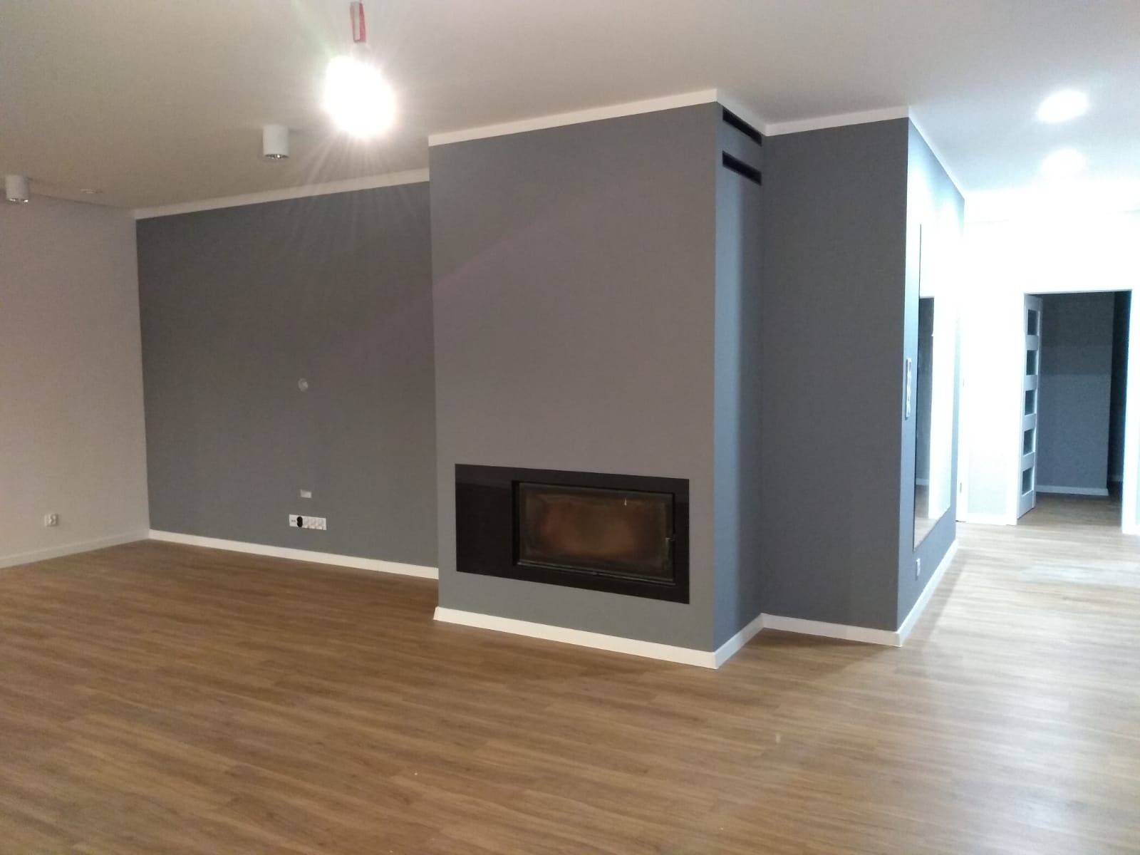 Szare ściany z kominkiem i drewnianą podłogą w pustym pokoju. Białe listwy przypodłogowe i sufitowe. Widok na korytarz z białymi drzwiami. Nowoczesne wykończenie.