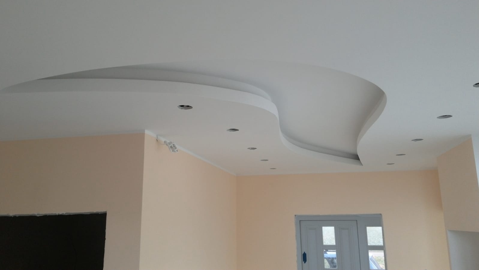 Nowoczesny sufit podwieszany z falistym kształtem i punktowym oświetleniem w jasnym wnętrzu. Ściany w odcieniach beżu, widoczne okno i otwór drzwiowy.
