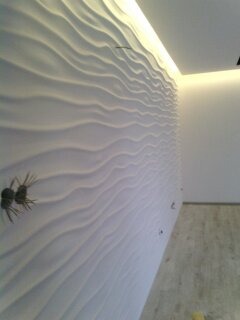 Biała ściana z dekoracyjnymi panelami 3D o falistym wzorze, oświetlona listwą LED przy suficie. Widoczne gniazdka elektryczne i fragment podłogi.