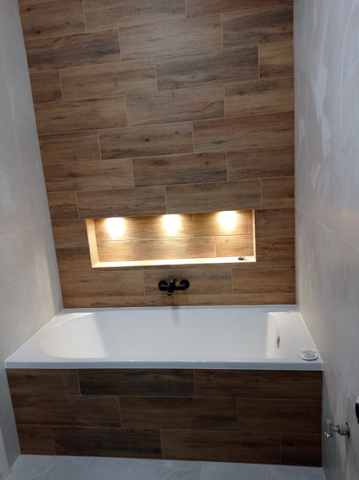 Nowoczesna łazienka z wanną obudowaną płytkami imitującymi drewno. W ścianie wnęka z oświetleniem punktowym. Czarna bateria wannowa. Minimalistyczny design.