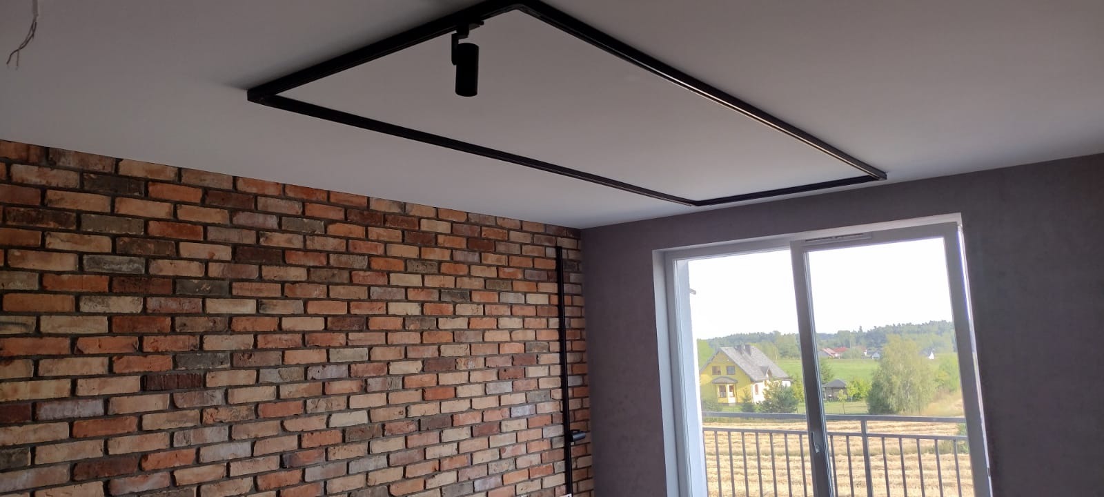 Wykończone wnętrze z ceglaną ścianą, oknem z widokiem na zewnątrz i nowoczesnym oświetleniem sufitowym z czarną ramą i reflektorem punktowym.