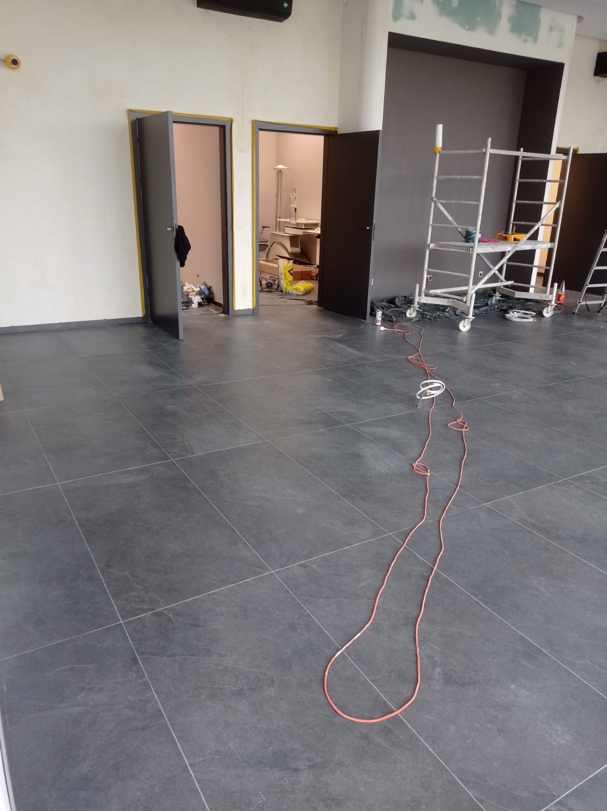Wnętrze w trakcie remontu: szare płytki podłogowe, otwarte drzwi, rusztowanie malarskie i przewód elektryczny na podłodze. Widoczne ślady prac wykończeniowych.