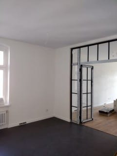 Białe ściany i czarna podłoga w pokoju z otwartymi, przeszklonymi drzwiami w czarnej ramie, prowadzącymi do jasnego pomieszczenia z drewnianą podłogą.