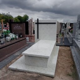 GALAN PRZEMYSŁAW ŁUKASZEWSKI - Jasny, nowoczesny nagrobek z czarnym krzyżem na tle pochmurnego nieba. Widoczne inne nagrobki i elementy cmentarza. Minimalistyczny design.
