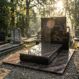 GALAN PRZEMYSŁAW ŁUKASZEWSKI - Nowoczesny nagrobek z ciemnego granitu z elementami w odcieniach brązu, oświetlony promieniami słońca na tle cmentarza z drzewami i innymi nagrobkami.