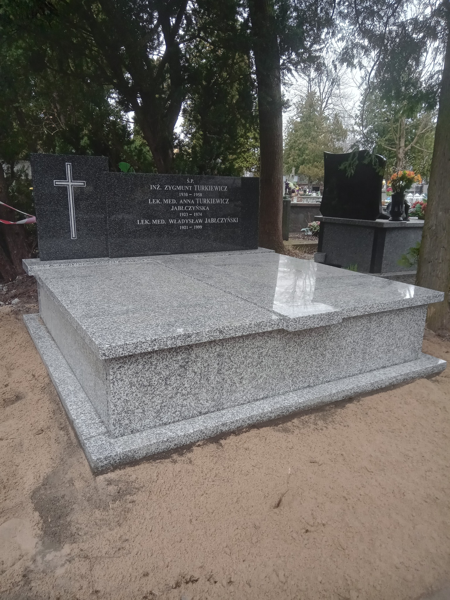 Granitowy nagrobek z krzyżem i inskrypcjami na cmentarzu, otoczony piaskiem. Widoczne inne nagrobki w tle, drzewa. Nagrobek w jasnych odcieniach szarości, wypolerowany.
