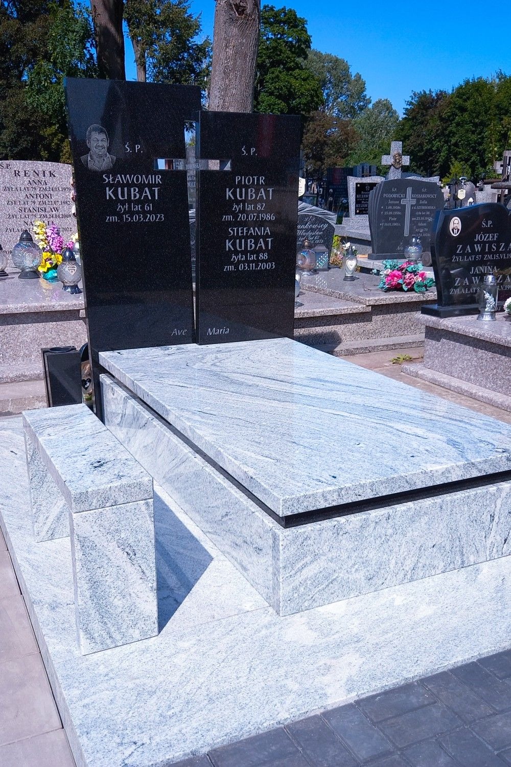 Granitowy nagrobek rodzinny z czarnymi tablicami i jasnoszarym obramowaniem, na tle cmentarza z drzewami i innymi nagrobkami w słoneczny dzień.