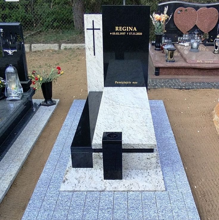 Czarno-biały nagrobek z krzyżem i inskrypcją 'Regina' na cmentarzu, otoczony innymi nagrobkami i kwiatami. Widok z przodu, oświetlenie dzienne.