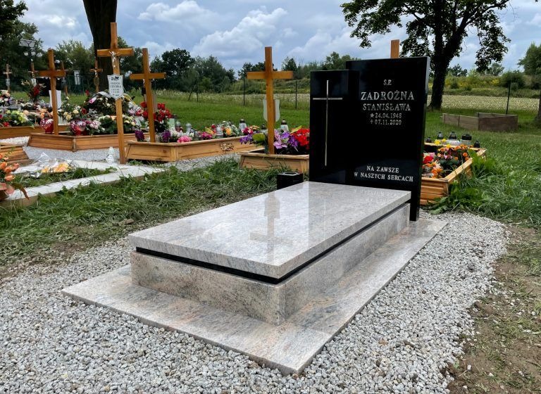 Granitowy nagrobek z czarną tablicą inskrypcyjną na cmentarzu, otoczony zielenią i innymi grobami. Jasny kamień nagrobka odbija światło.