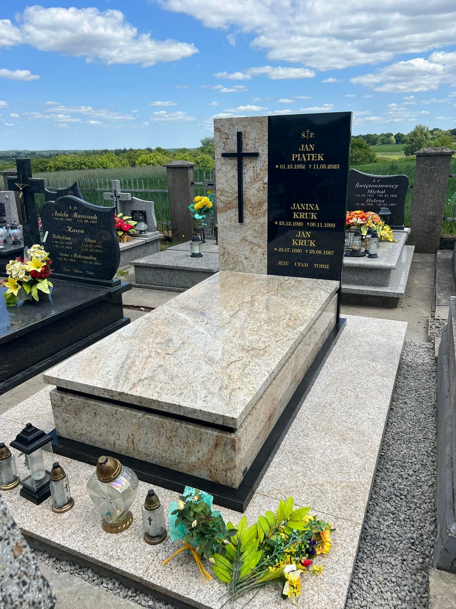 Jasny nagrobek z granitu z czarną tablicą i krzyżem, otoczony kwiatami i zniczami na cmentarzu w słoneczny dzień. Widok na zielone pola w tle.