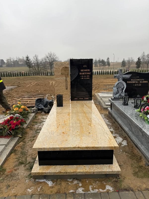 Nowy, jasny nagrobek z ciemnymi elementami na cmentarzu, z widocznymi nagrobkami w tle. Na płycie nagrobnej wyryte imię i nazwisko oraz daty urodzin i śmierci.