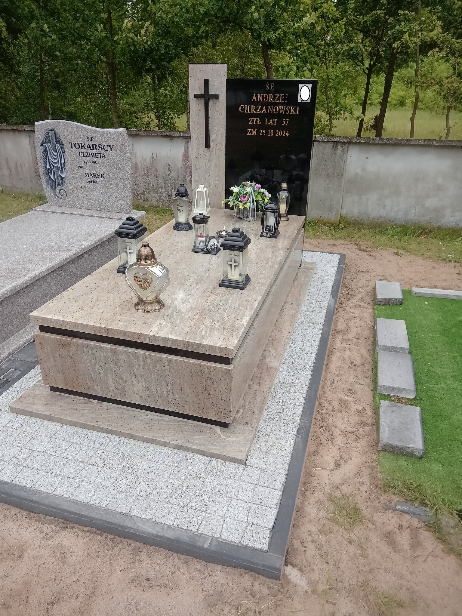 Jasny nagrobek z beżowego kamienia z czarną tablicą i krzyżem, otoczony szarą kostką brukową i elementami trawy. Widoczne znicze i kwiaty.