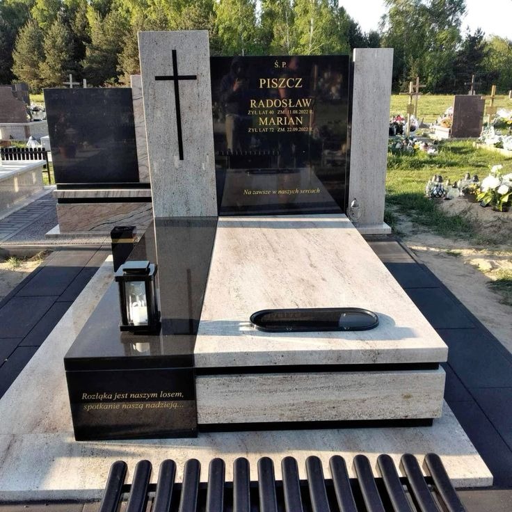 Elegancki, dwukolorowy nagrobek z czarnego i jasnego kamienia, z wyrytym krzyżem i inskrypcjami, otoczony zielenią cmentarza. Widoczna ławka przed pomnikiem.