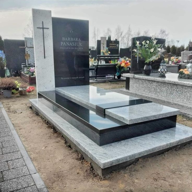 Elegancki, dwukolorowy nagrobek z jasnego i czarnego granitu, z wyrytym krzyżem i inskrypcją, na tle cmentarza z innymi nagrobkami i zielenią.