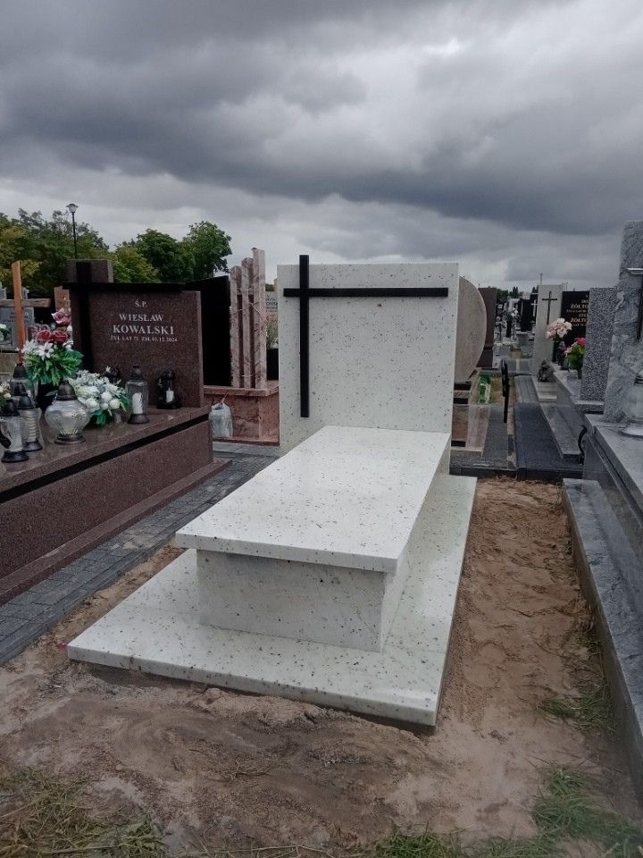 Jasny, nowoczesny nagrobek z czarnym krzyżem na tle pochmurnego nieba. Widoczne inne nagrobki i elementy cmentarza. Minimalistyczny design.