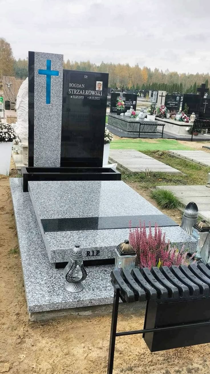 Granitowy nagrobek z niebieskim krzyżem na cmentarzu, z inskrypcją i zdjęciem zmarłego, otoczony kwiatami i lampionami, na tle innych grobów i drzew.